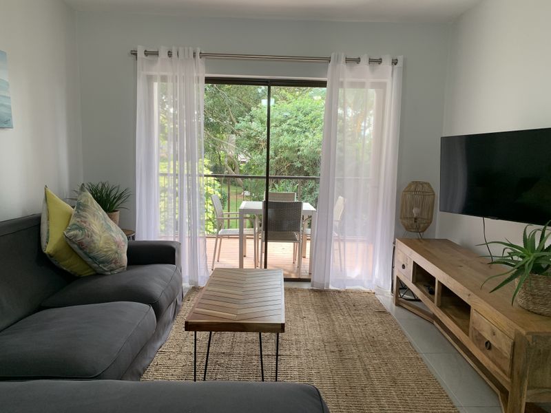 A bright living room featuring tiled floors and a large sliding glass door that opens onto a private balcony with garden views.
