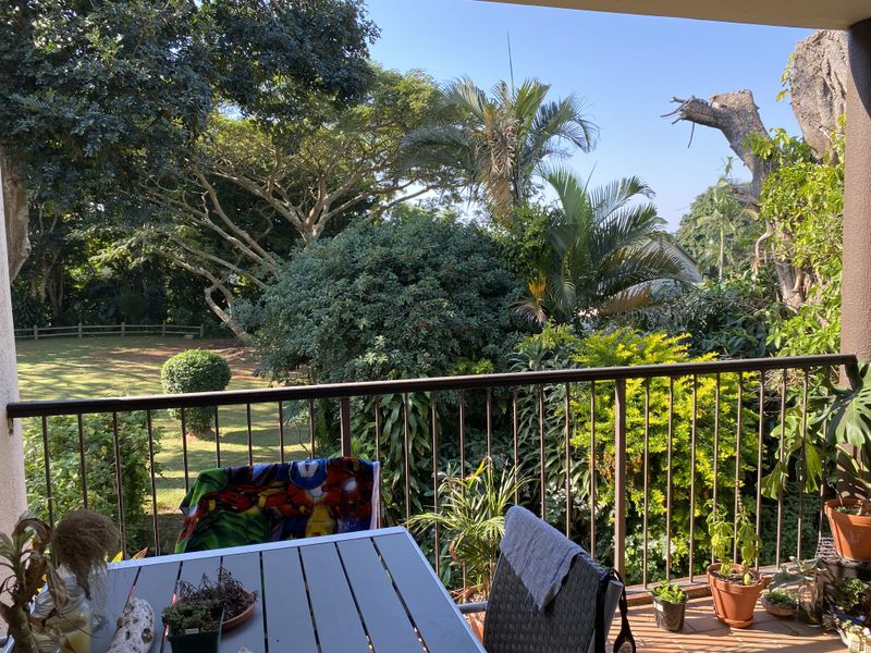 A private balcony featuring a metal balustrade and tiled flooring, overlooking a lush, mature garden with established trees.