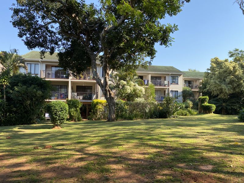 A view of the rear elevation of the apartment complex, featuring private balconies and a well-maintained, spacious communal garden with mature trees.
