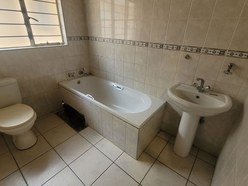 A dated bathroom featuring a bathtub, pedestal basin, and toilet with neutral wall and floor tiling.