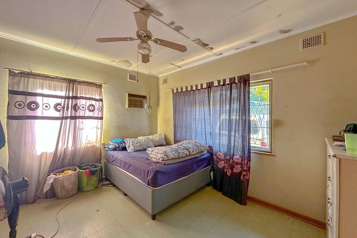 A bedroom featuring a ceiling fan, wall-mounted air conditioning unit, and dual windows, currently showing signs of wear and ceiling water damage.