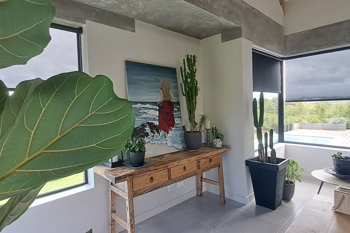 Modern living area featuring exposed concrete beams, large format floor tiles, and expansive black-framed windows with roller blinds, offering a bright and contemporary aesthetic.