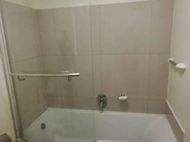 A functional bathroom featuring a built-in bathtub with a glass shower screen, chrome mixer tap, and neutral wall tiling.