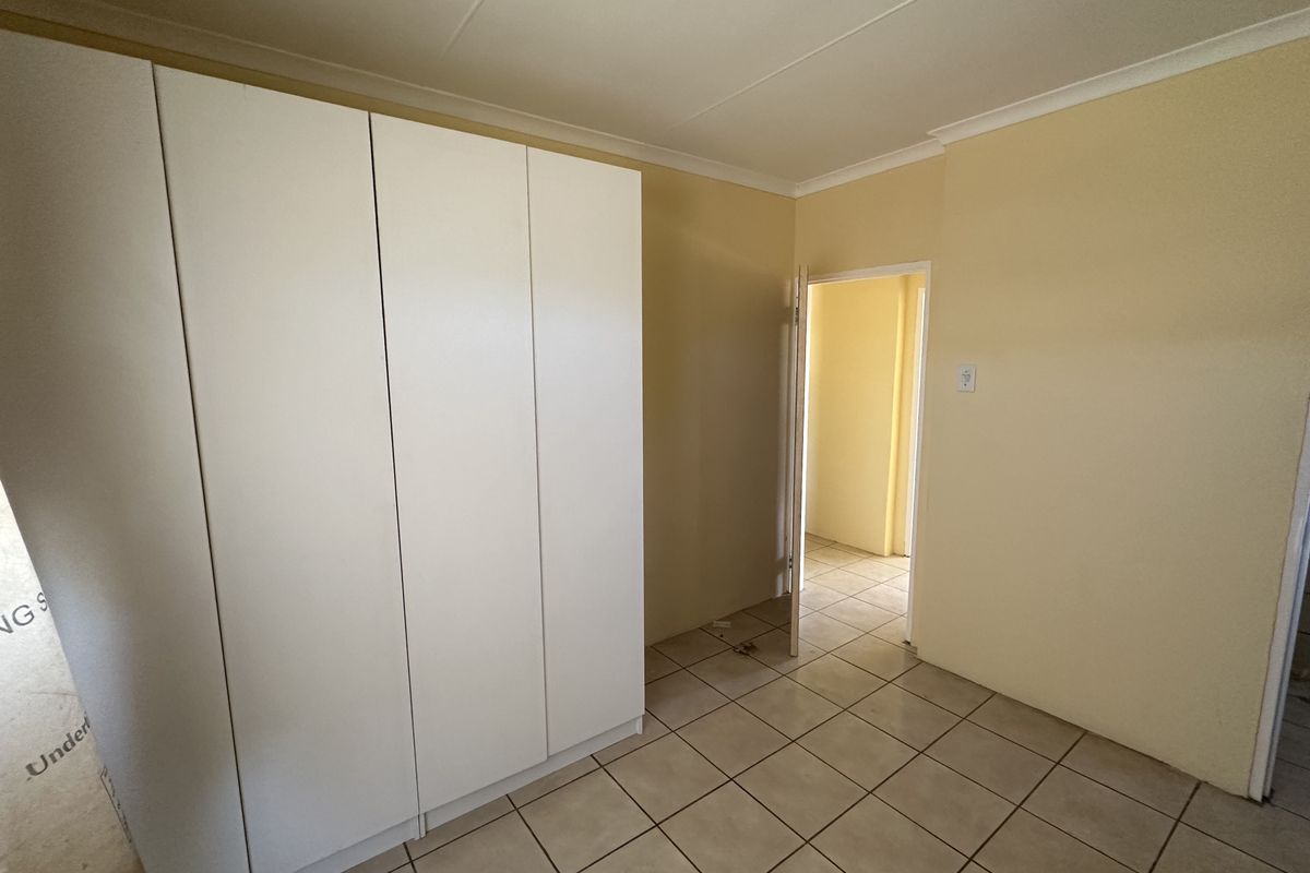 A clean, empty bedroom featuring neutral tiled flooring, a built-in white wardrobe, and access to an adjoining hallway.