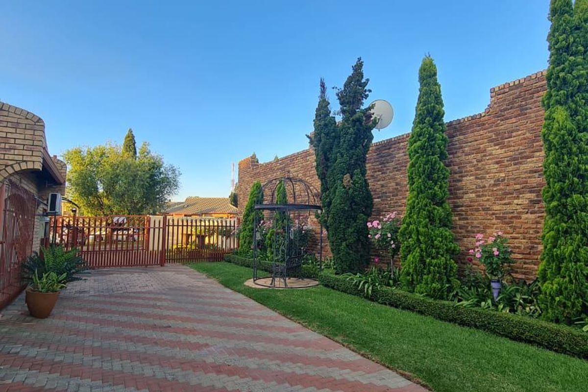 A well-maintained outdoor area featuring a paved driveway, manicured lawn, established cypress trees, and a high brick boundary wall.