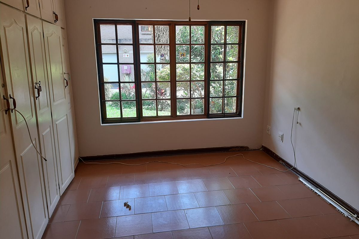 An empty bedroom featuring tiled flooring, built-in cupboards, and large wooden-framed windows overlooking the garden.