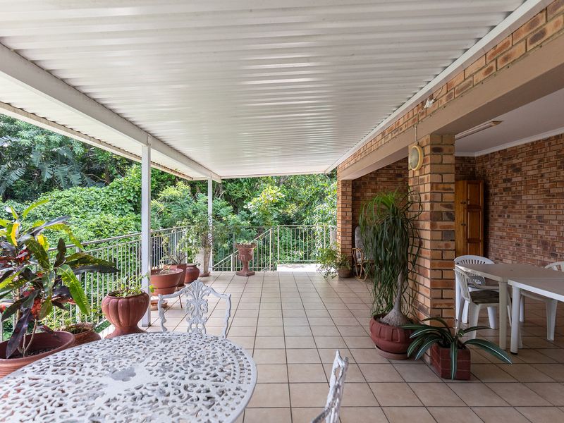Spacious covered patio featuring brick pillars, tiled flooring, and metal railings, overlooking a lush garden area.