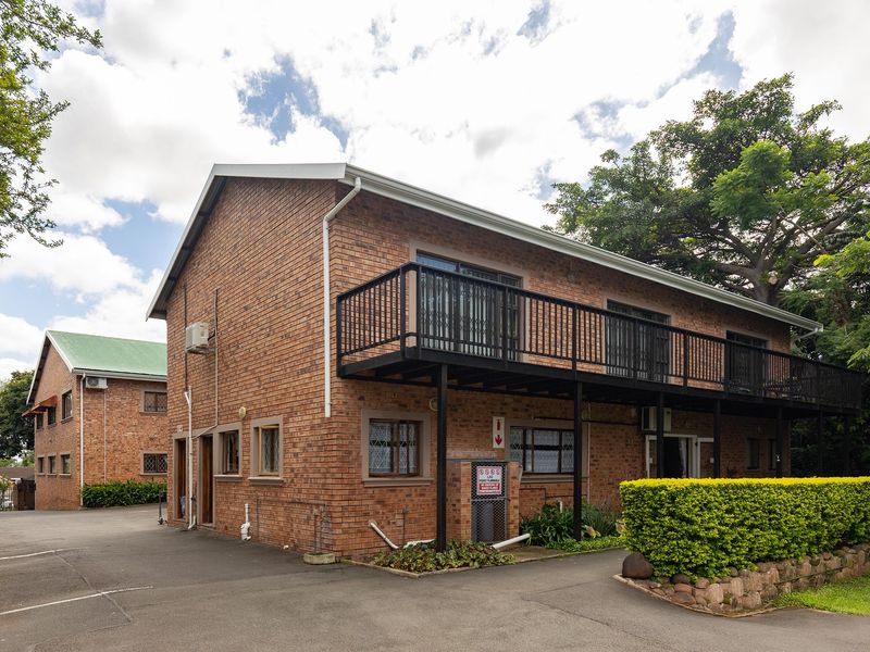 Exterior view of a two-story face brick retirement village building featuring a raised metal balcony, paved driveway, and established garden landscaping.
