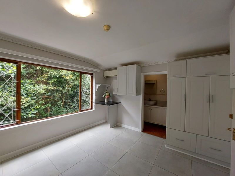 A bright, empty bedroom featuring tiled floors, built-in cabinetry, decorative crown molding, and a large window overlooking lush greenery, with an adjoining bathroom.