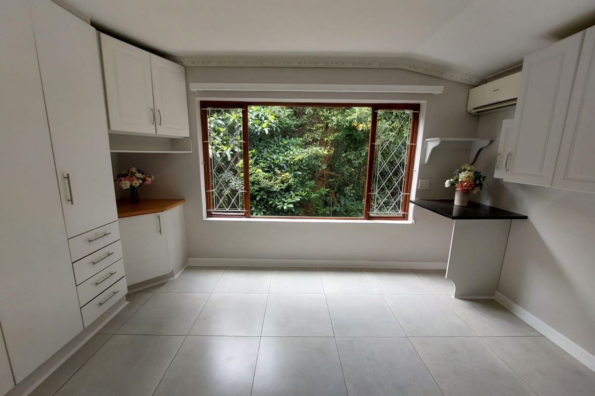 A bright, empty kitchen featuring white built-in cabinetry, tiled flooring, and a large window overlooking lush greenery, offering a clean and functional space.
