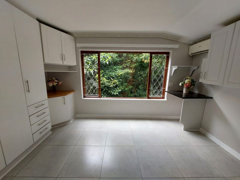 A bright, empty kitchen featuring white built-in cabinetry, tiled flooring, and a large window overlooking lush greenery, offering a clean and functional space.