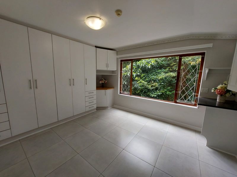 A bright, empty bedroom featuring extensive built-in white cabinetry, large windows with security bars, and neutral tiled flooring.