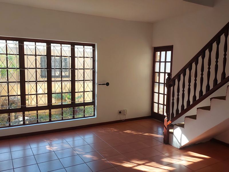 An empty living area featuring tiled flooring, a wooden staircase with a balustrade, and large windows fitted with security bars.