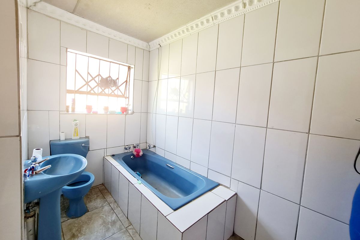 A dated bathroom featuring blue sanitaryware, full-height white wall tiling, and tiled flooring with security bars on the window.