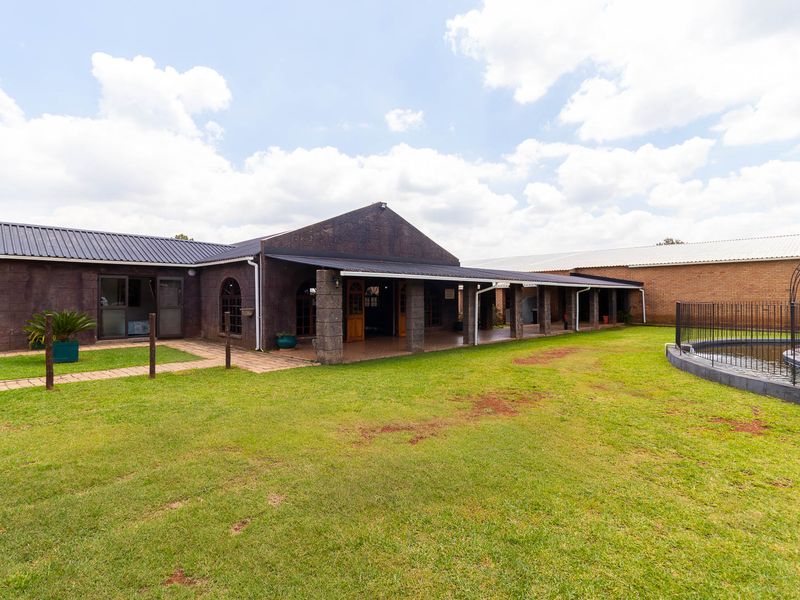 Rear view of the farm property featuring a spacious covered patio, stone-clad exterior, and a fenced swimming pool set within a large lawn area.