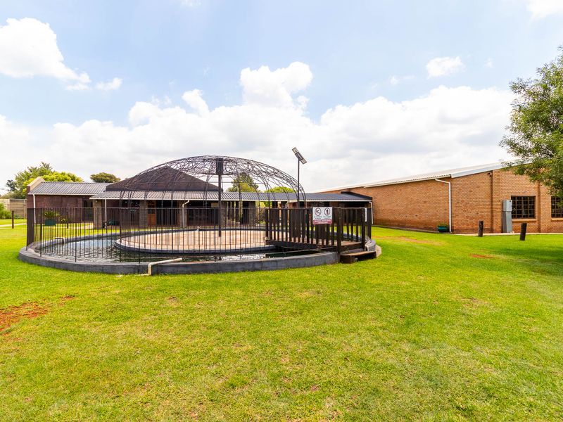 A landscaped garden area featuring a circular water feature with a decorative metal dome structure, surrounded by lawn and brick farm buildings.
