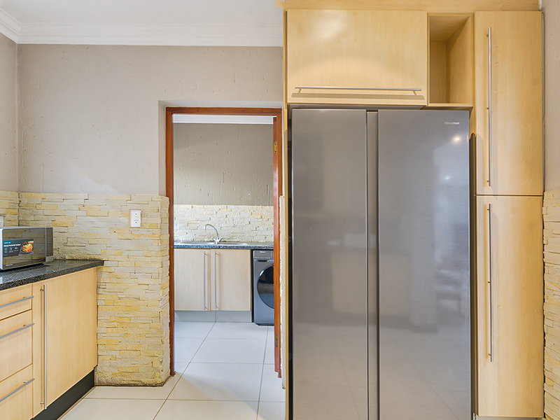 A functional kitchen featuring light wood cabinetry, granite countertops, and a decorative stone backsplash, with an adjacent scullery area visible through the doorway.