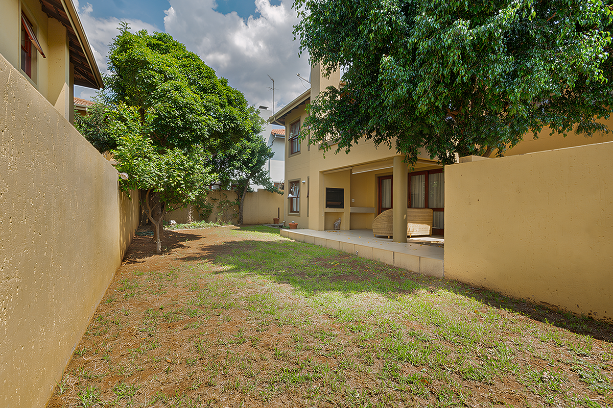 A private rear garden featuring a covered patio with a built-in braai, enclosed by high perimeter walls and mature trees.