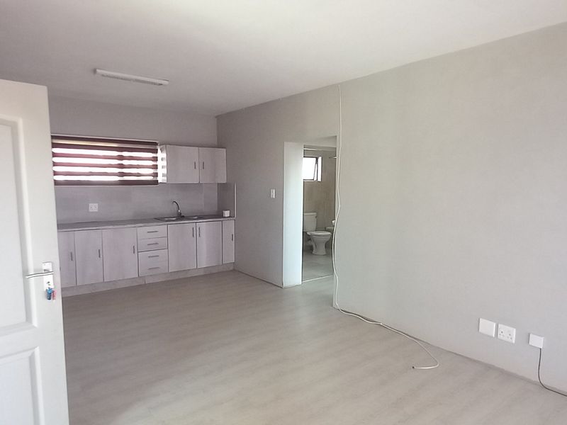 An open-plan living area featuring light-toned laminate flooring, a compact kitchen with built-in cabinetry, and direct access to an adjacent bathroom.
