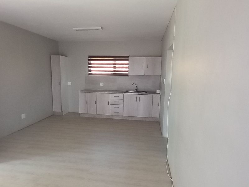 An open-plan living and kitchen area featuring light-toned laminate flooring, neutral walls, and functional kitchen cabinetry with a built-in sink.