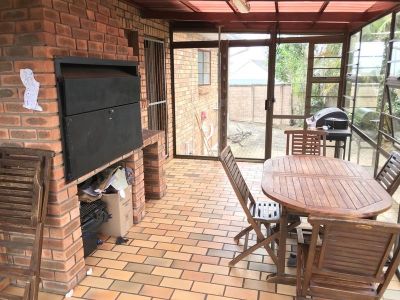 A covered patio area featuring a built-in brick braai, brick flooring, and large windows overlooking the rear garden.