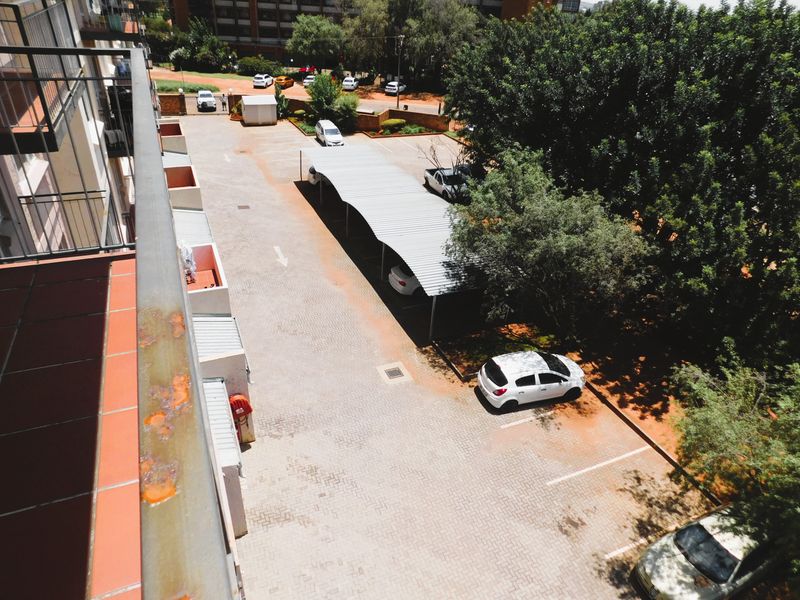 Elevated view of the apartment complex parking area featuring paved surfaces and covered carports, viewed from a balcony with a metal railing.