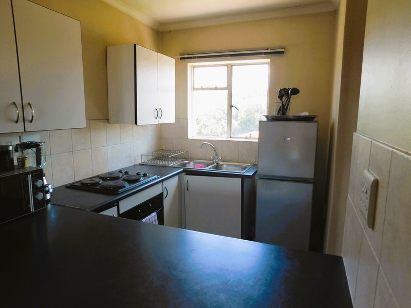 A compact kitchen featuring white cabinetry, dark laminate countertops, a built-in electric hob and oven, and a stainless steel sink with a tiled backsplash.