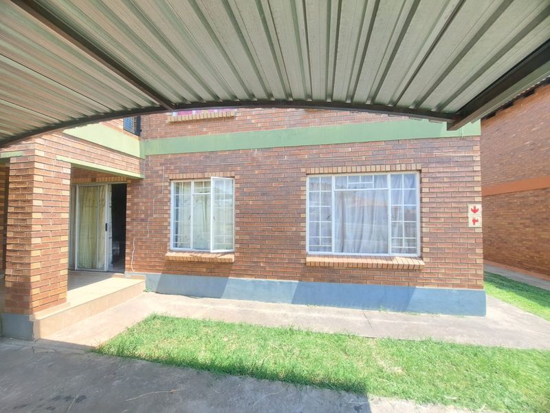 Exterior view of a face brick apartment building featuring a metal carport, steel-framed windows, and a paved walkway entrance.