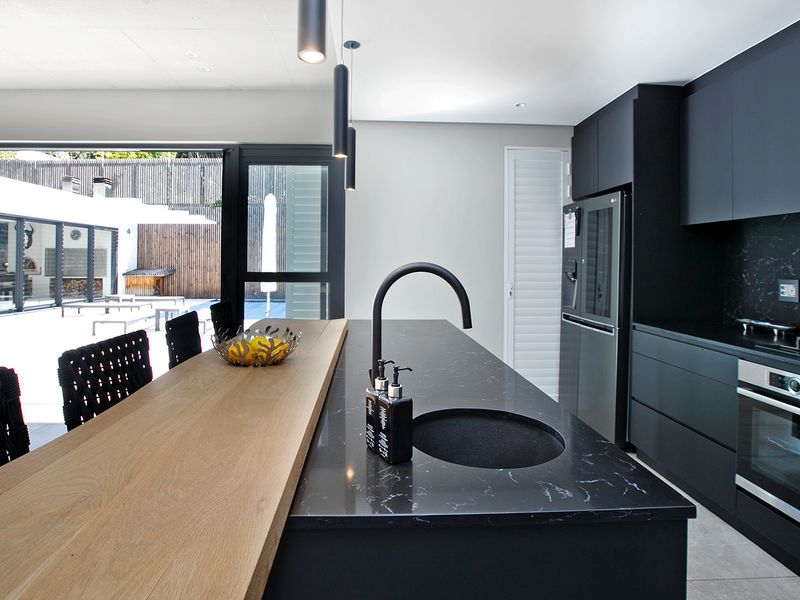 Modern open-plan kitchen featuring a sleek black island with integrated sink, wooden breakfast bar, and high-end built-in appliances, seamlessly connecting to the outdoor patio.