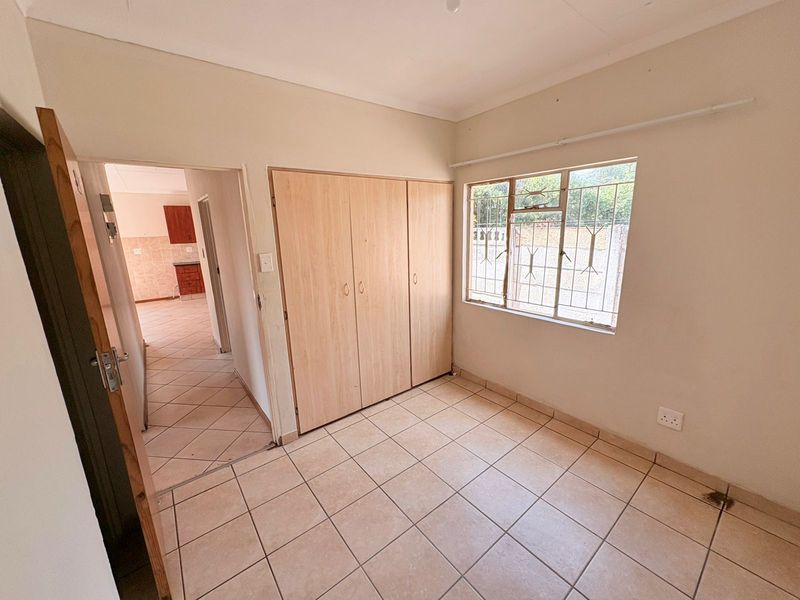 A vacant bedroom featuring built-in cupboards, tiled flooring, and security bars on the window, with a view into the adjacent kitchen area.