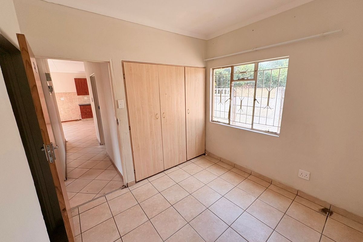A vacant bedroom featuring built-in cupboards, tiled flooring, and security bars on the window, with a view into the adjacent kitchen area.