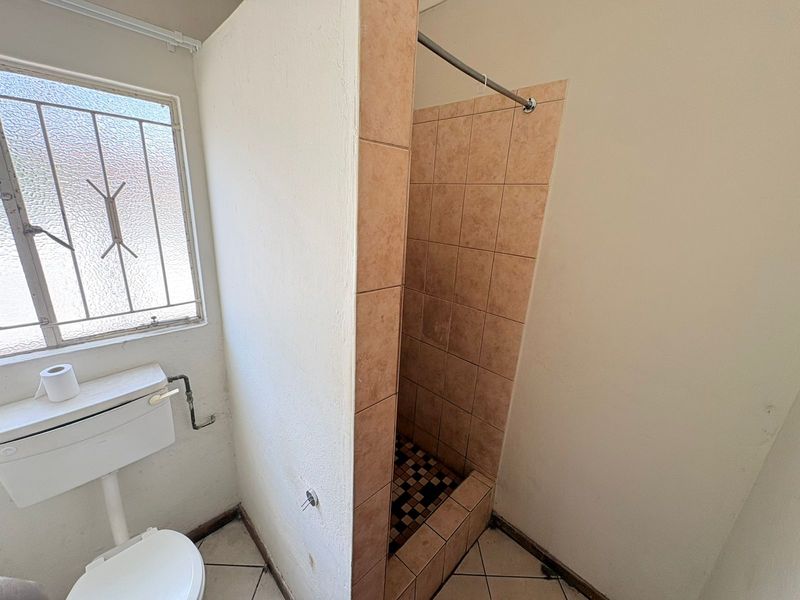 A functional bathroom featuring a tiled shower cubicle, toilet, and security-barred window. The space requires cosmetic updates to modernize the dated finishes.