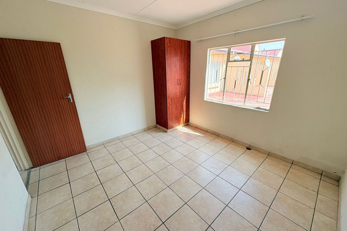 A simple, empty bedroom featuring tiled flooring, a built-in wardrobe, and a window fitted with security bars.