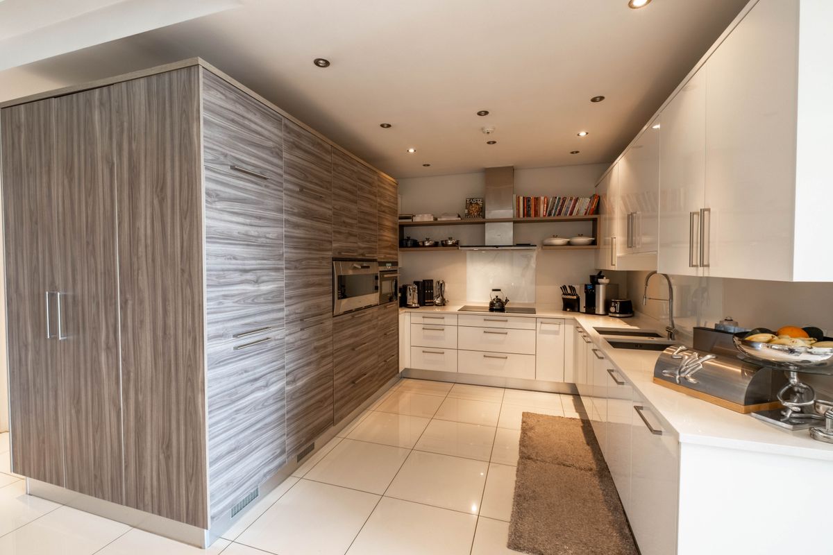 Modern kitchen featuring high-gloss white cabinetry, wood-grain integrated storage, built-in oven, microwave, extractor fan, and sleek tiled flooring.