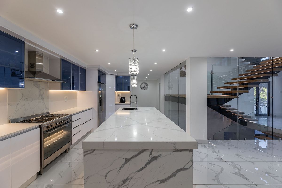 Modern kitchen featuring a marble-look quartz island, gas stove, high-gloss cabinetry, and an adjacent floating staircase with glass balustrades.