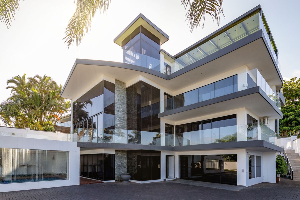 A modern multi-level architectural home featuring extensive glass facades, stone cladding, glass balustrades, and a paved driveway with an integrated water feature.