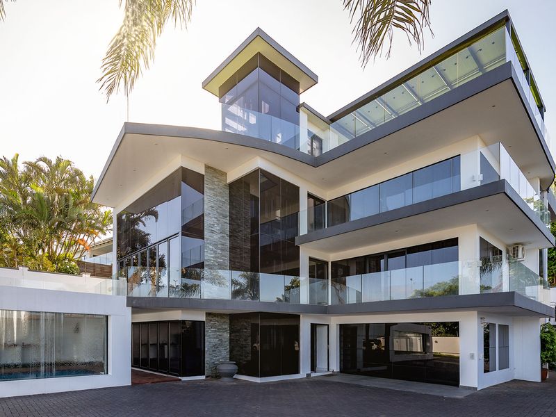 A modern multi-level architectural home featuring extensive glass facades, stone cladding, glass balustrades, and a paved driveway with an integrated water feature.