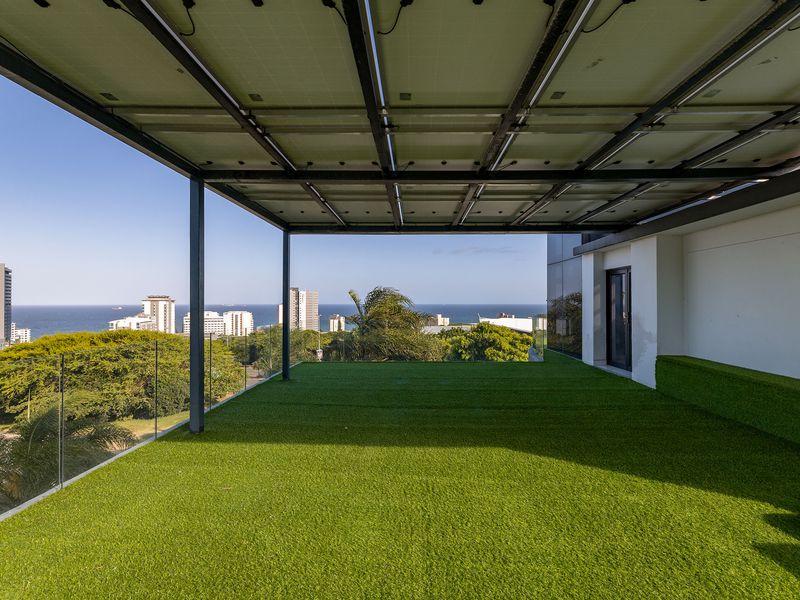 Spacious covered balcony featuring artificial turf, a glass balustrade, and integrated solar panels, offering expansive coastal and city views.
