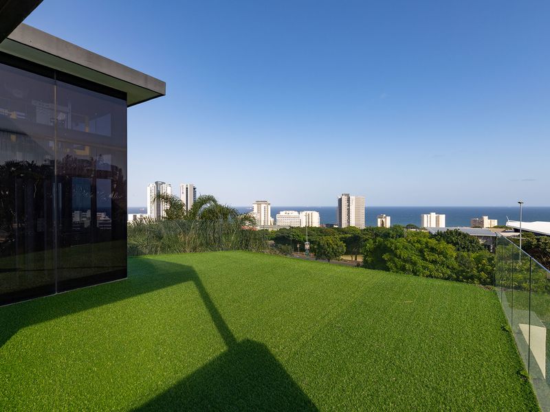 A modern elevated balcony featuring artificial turf, a sleek glass balustrade, and expansive views of the city skyline and ocean.