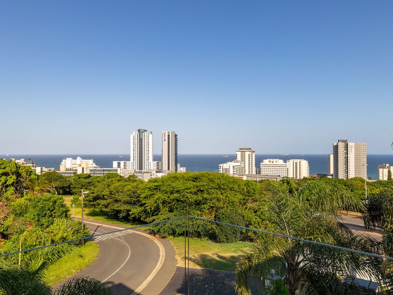 Elevated panoramic view overlooking the surrounding neighborhood, lush greenery, and the distant city skyline against the ocean horizon.