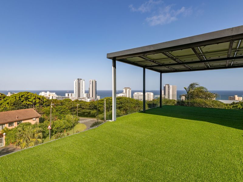 Spacious elevated balcony featuring artificial turf, a modern metal pergola, and a glass balustrade with panoramic ocean and city views.
