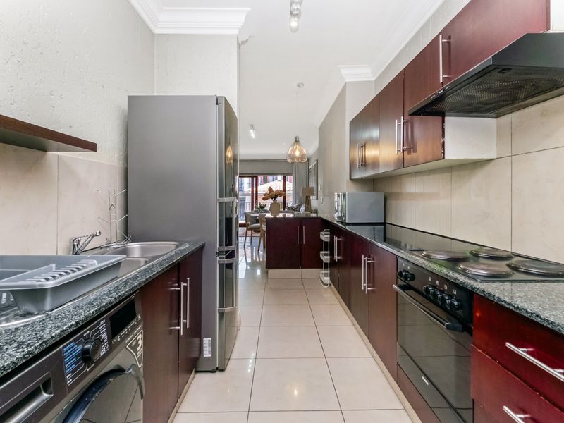 A functional galley-style kitchen featuring dark wood cabinetry, granite countertops, built-in oven, electric hob, and tiled flooring, leading into an open-plan living area.