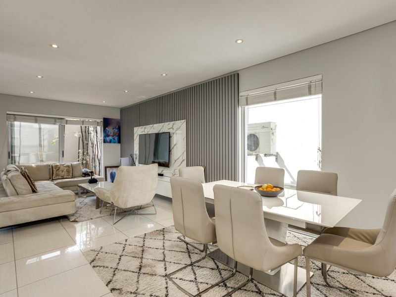 A bright open-plan living and dining area featuring polished floor tiles, recessed downlighting, and a modern feature wall with integrated television mounting.