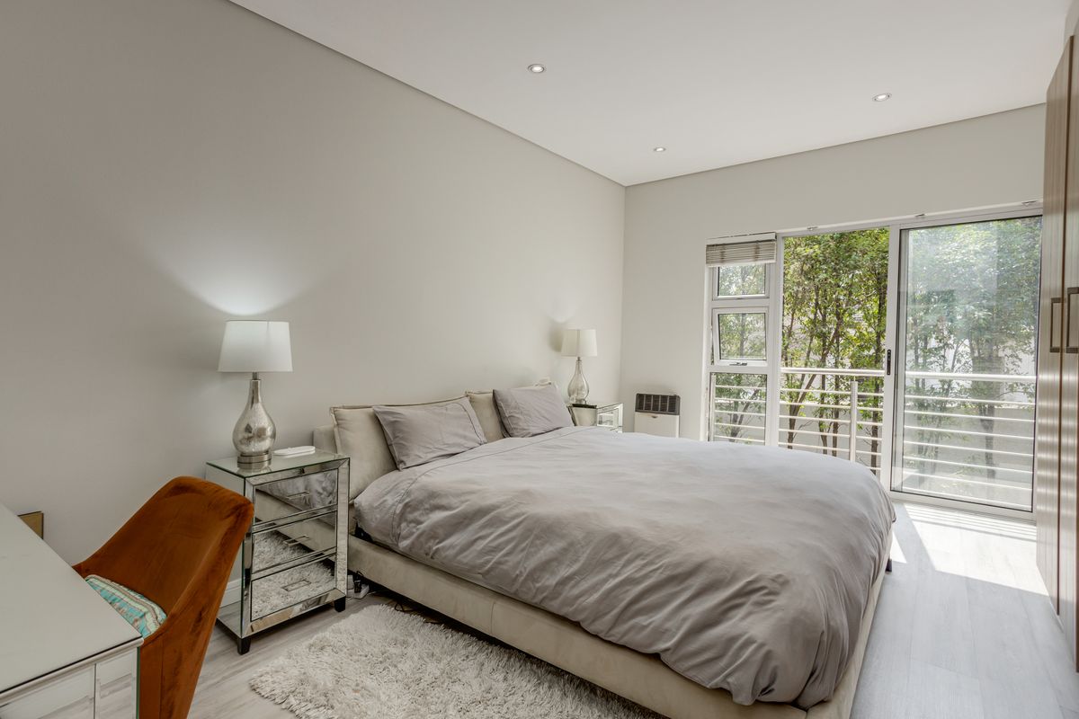 A bright and modern bedroom featuring built-in wardrobes, recessed ceiling lighting, and large sliding glass doors that open to a balcony with leafy views.
