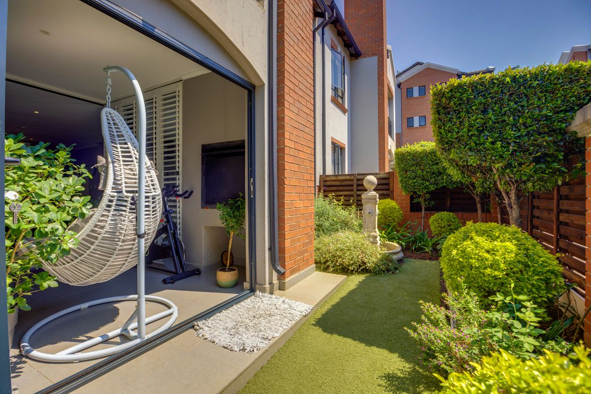 A private rear garden featuring a manicured lawn, established shrubbery, a decorative stone fountain, and a covered patio area with a built-in braai.