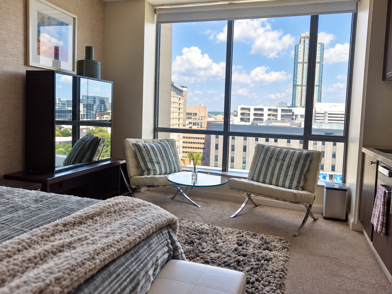 A modern open-plan studio apartment featuring floor-to-ceiling windows with city views, carpeted flooring, and a compact kitchenette with built-in cabinetry.