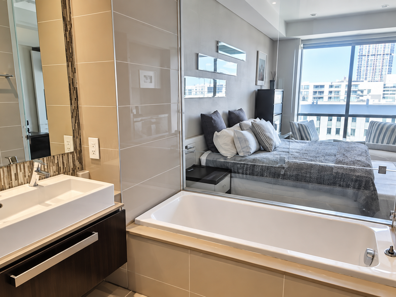 Modern bathroom featuring a sleek vanity, built-in bathtub, and a glass partition wall overlooking the adjacent bedroom with city views.