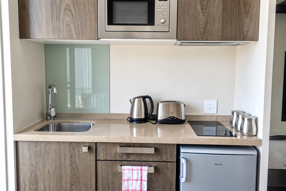 Compact modern kitchen featuring wood-grain cabinetry, stone countertops, integrated microwave, electric cooktop, and a stainless steel sink with a glass splashback.