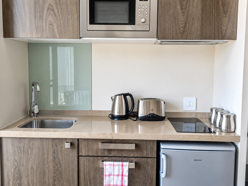 Compact modern kitchen featuring wood-grain cabinetry, stone countertops, integrated microwave, electric cooktop, and a stainless steel sink with a glass splashback.