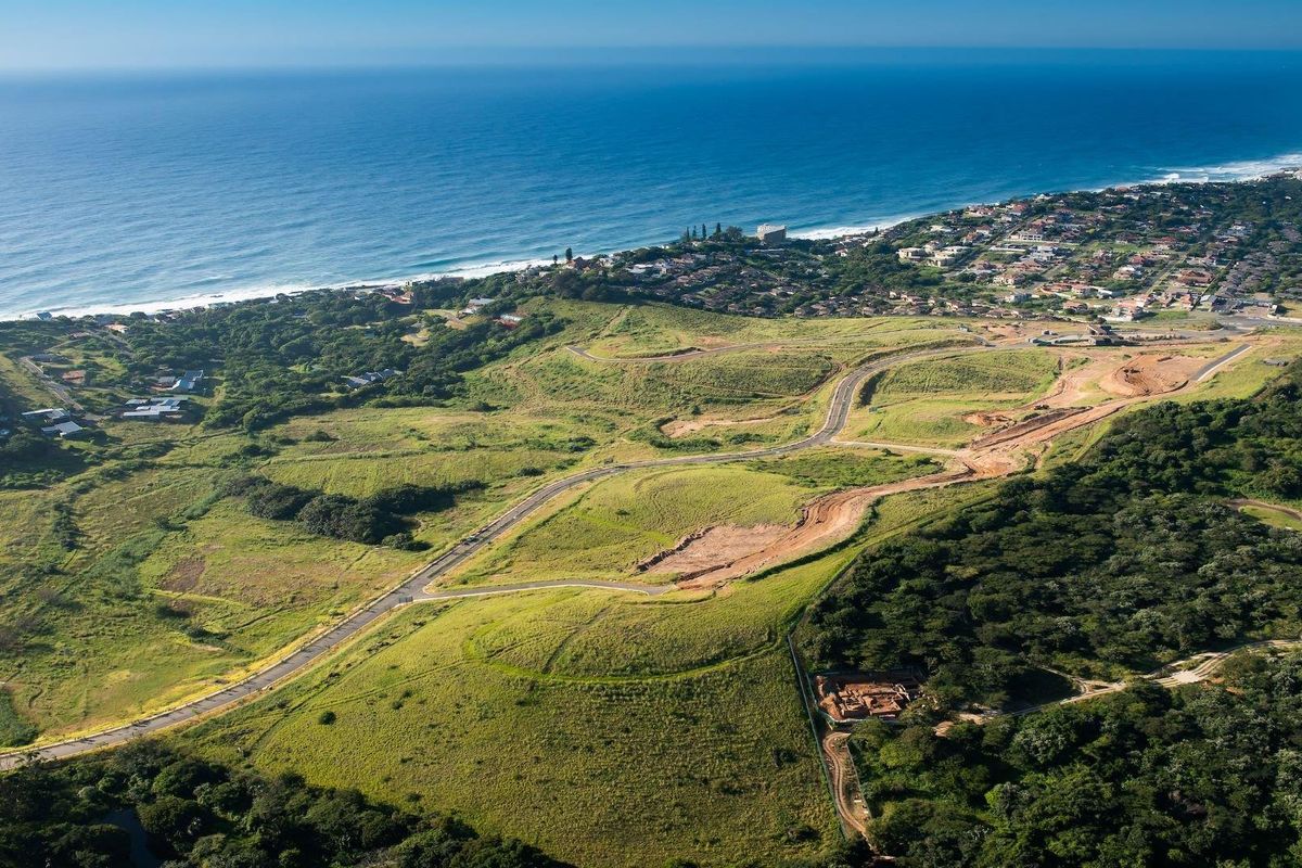 An aerial view of a coastal development site featuring established road infrastructure, surrounding greenery, and proximity to the ocean.